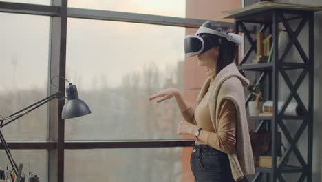 Portrait.-Female-architect-designer-in-a-virtual-reality-helmet-at-in-a-modern-office-near-a-large-window-with-his-hands-imitates-the-work-of-interface.-Designing-the-future-the-concept-of-virtual-architecture-and-design-interface-graphic-applications.
